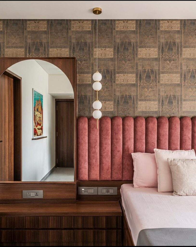 Elegant bedroom interior design by Olangana Design in Bangalore featuring a pink upholstered headboard, a stylish wooden dresser with an arched mirror, modern pendant lighting, and patterned wallpaper, showcasing a sophisticated residential space.