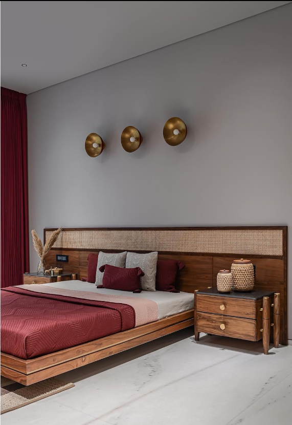 Modern bedroom interior designed by Olangana Design in Bangalore, featuring a wooden bed with rattan headboard, maroon bedding, and contemporary brass wall sconces.