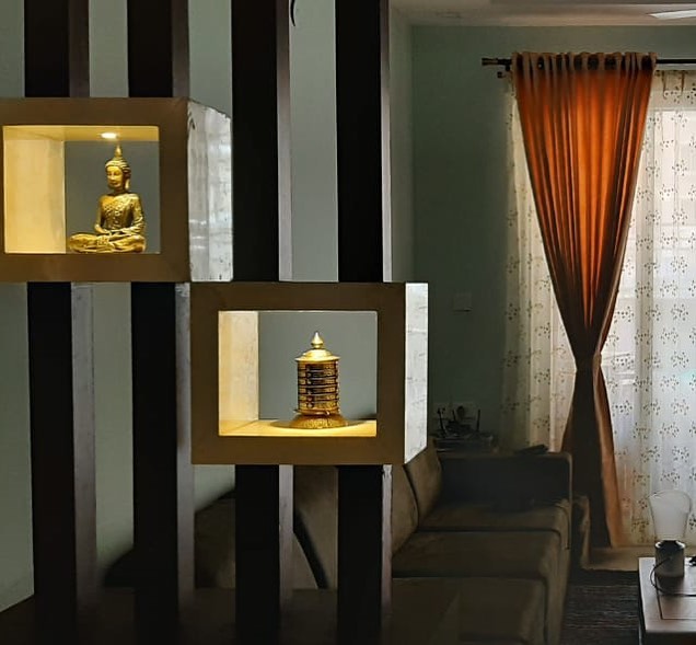 Modern living room interior design by Olangana Design in Bangalore featuring a decorative partition with illuminated niches showcasing a Buddha statue and a golden prayer wheel.