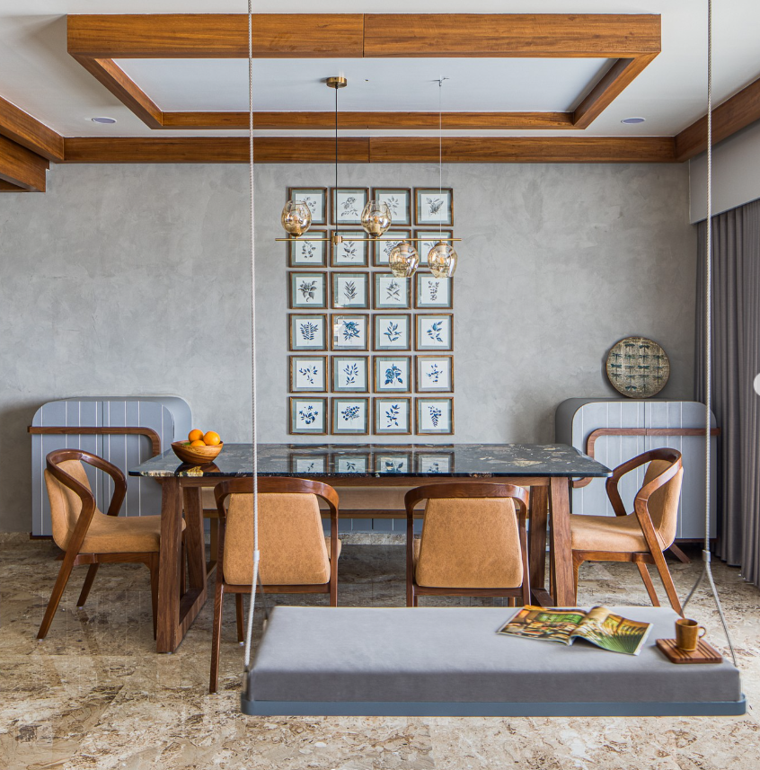 Modern dining room interior by Olangana Design in Bangalore, featuring a dark marble table, wooden chairs, a grey indoor swing, and a striking gallery wall with blue and white framed art.
