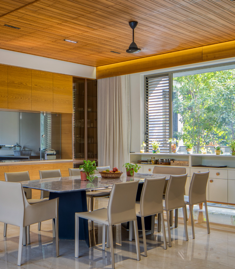 Modern dining room interior by Olangana Design in Bangalore featuring a sleek marble dining table, elegant beige chairs, and a warm wooden slatted ceiling with ample natural light.