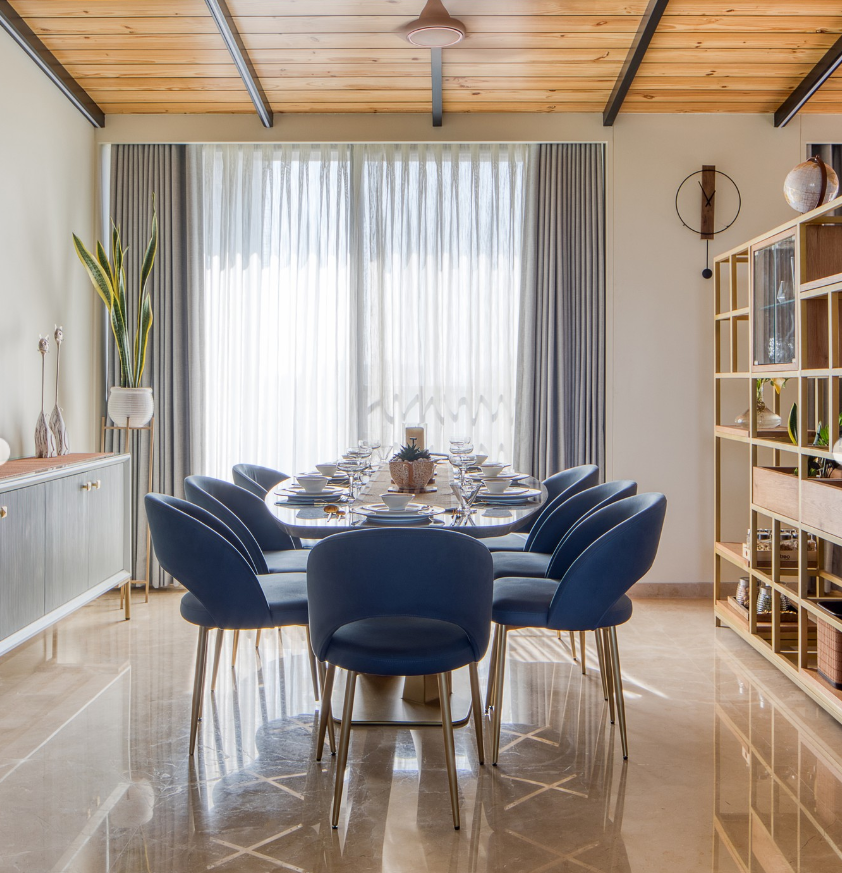 Modern dining room interior by Olangana Design in Bangalore featuring a sleek oval table, blue velvet chairs, and a unique wooden slatted ceiling design, embodying sophisticated interior craftsmanship.