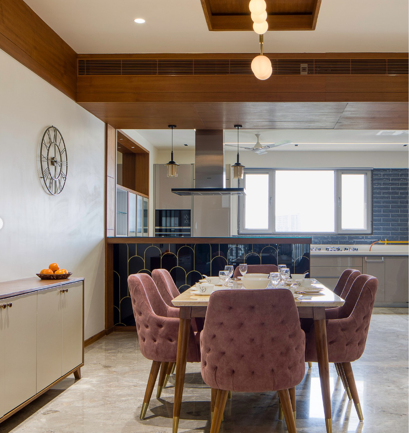 Modern dining room and open-concept kitchen interior design by Olangana Design in Bangalore, featuring a marble dining table, tufted pink chairs, and a stylish patterned bar counter leading to a sleek kitchen.