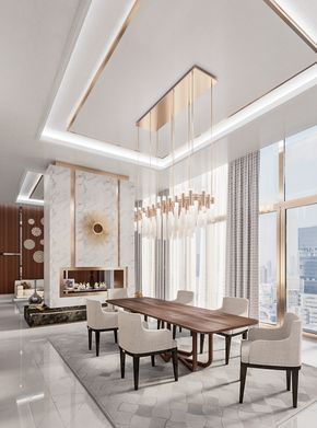 Luxury modern dining room interior design by Olangana Design in Bangalore, featuring a long wooden dining table, upholstered chairs, a striking gold chandelier, and a marble fireplace with a golden sunburst accent. Large windows offer a city view.