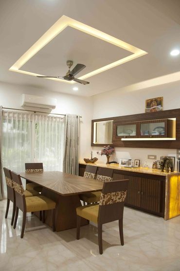 Elegant modern dining room interior design by Olangana Design in Bangalore, featuring a stylish wooden dining table, upholstered chairs, a decorative false ceiling with cove lighting, and a sophisticated buffet unit with backlit onyx.