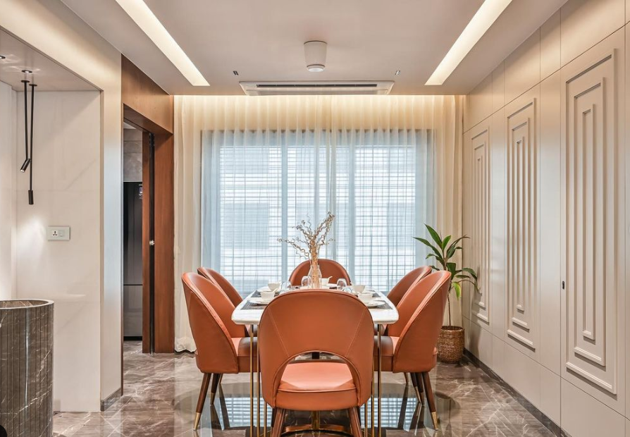 Elegant modern dining interior design by Olangana Design in Bangalore featuring burnt orange velvet chairs, a white marble dining table, and sophisticated lighting with decorative wall paneling.