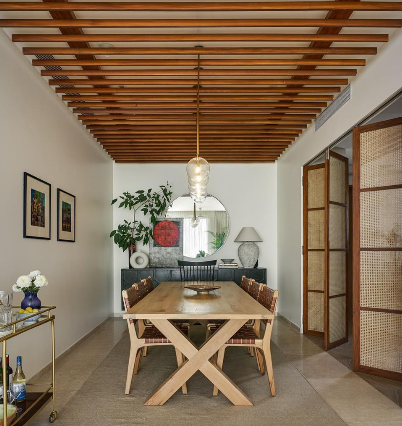 Modern dining room interior by Olangana Design in Bangalore, featuring a unique wooden slatted ceiling, a large wooden dining table, contemporary chairs, a console table with decor, and rattan bi-fold doors.