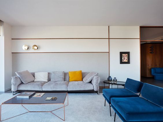 Modern living room interior design by Olangana in Bangalore, featuring a grey velvet sofa, vibrant blue armchairs, a geometric copper coffee table, and elegant wall paneling with gold sconces.