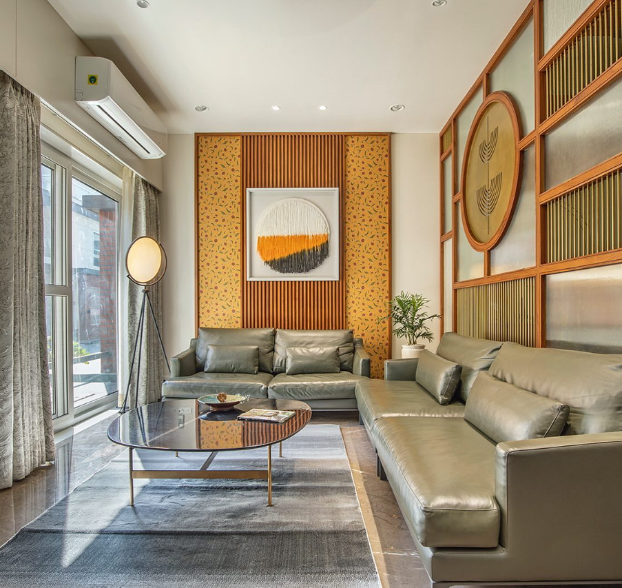 Modern living room interior design by Olangana in Bangalore, featuring elegant olive green leather sofas, a sleek dark coffee table, and distinctive wall decor with wooden slats, patterned wallpaper, and brass art panels.