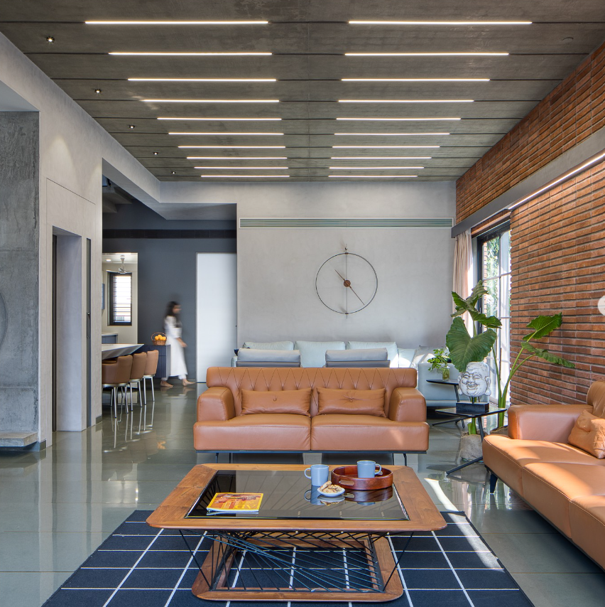 Modern living room interior design by Olangana in Bangalore, featuring warm brown leather sofas, an exposed brick accent wall, and a concrete ceiling with linear lighting.