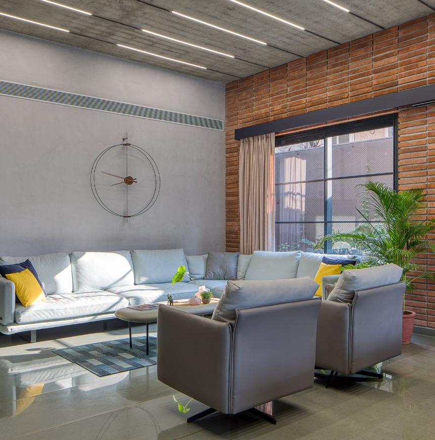 Modern industrial living room interior design by Olangana in Bangalore, featuring a light grey L-shaped sofa, exposed brick wall, concrete ceiling with linear lighting, and a minimalist wall clock.