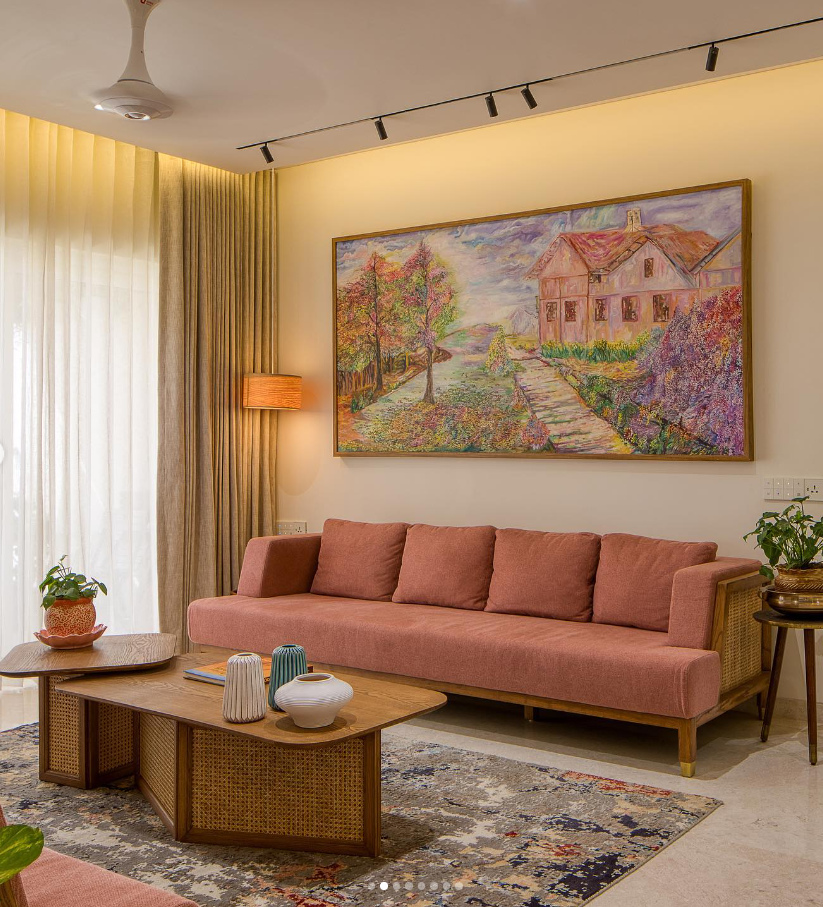 Modern living room interior design by Olangana in Bangalore, featuring a salmon-colored sofa with rattan details, elegant wooden coffee tables, and a vibrant landscape painting, creating a warm and inviting atmosphere.