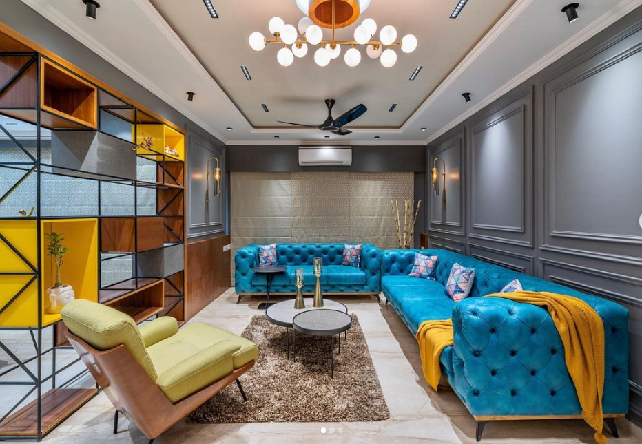 Modern living room interior design by Olangana in Bangalore, featuring a vibrant teal tufted L-shaped sofa, a striking yellow and wood open shelving unit, and elegant grey paneled walls.