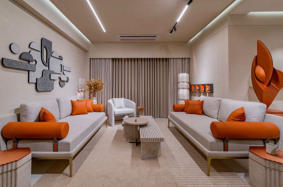 Elegant living room interior design by Olangana in Bangalore, featuring neutral sofas with vibrant orange cushions, abstract wall art, and modern ceiling lighting.