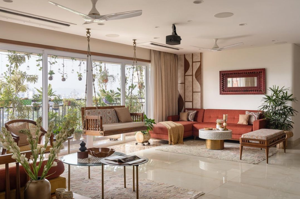 Elegant living room interior design by Olangana in Bangalore, featuring a warm terracotta sectional sofa, a traditional wooden swing, lush balcony greenery, and a carved mirror, creating a serene and inviting space.