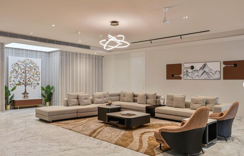 Modern living room interior design by Olangana in Bangalore, featuring a large neutral sectional sofa, elegant accent chairs, a striking circular chandelier, and unique wall art including a tree of life and abstract mountains.