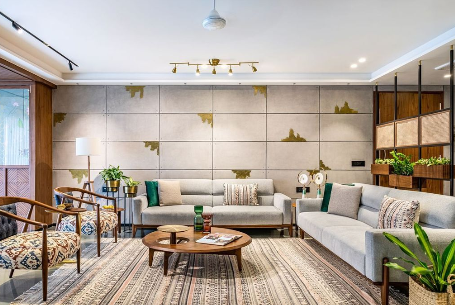 Modern living room interior design by Olangana in Bangalore, featuring sleek grey sofas, a round wooden coffee table, unique concrete wall panels with abstract gold accents, and a stylish patterned rug.