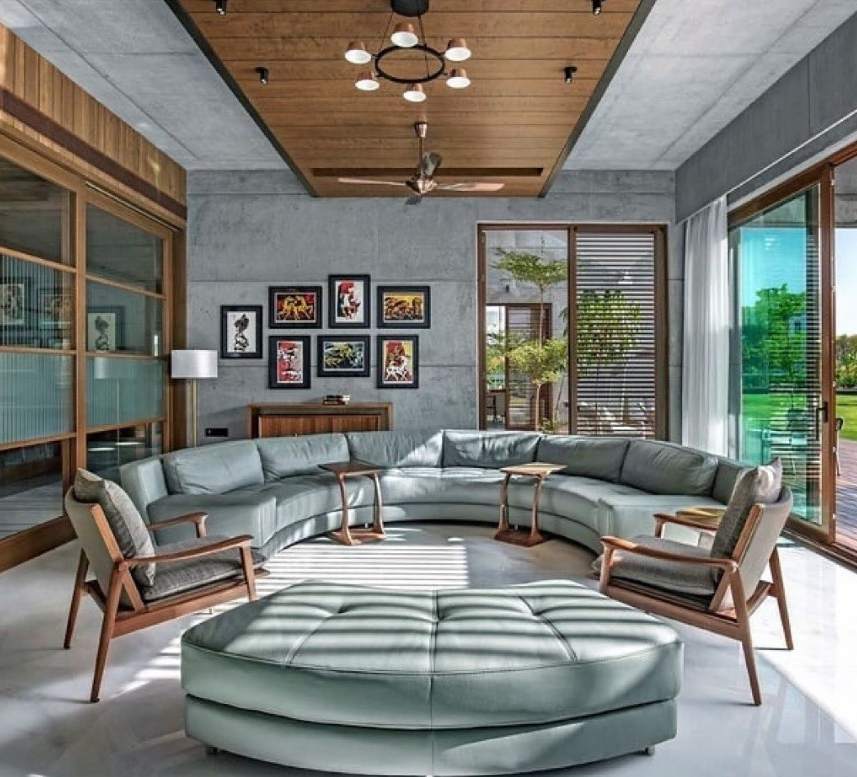 Mid-century modern living room interior design by Olangana in Bangalore, featuring warm wood paneling, a tan leather sofa, blue velvet armchairs, and vibrant abstract art.
