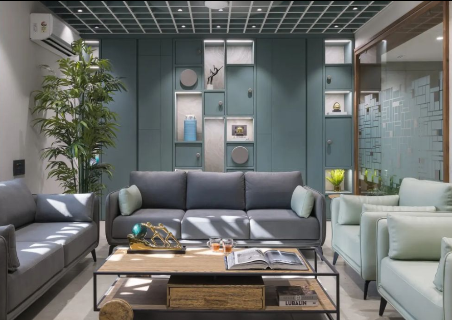 Modern living room interior design by Olangana in Bangalore, featuring a teal accent wall with integrated display shelves, comfortable grey and mint green sofas, and a stylish wooden coffee table.