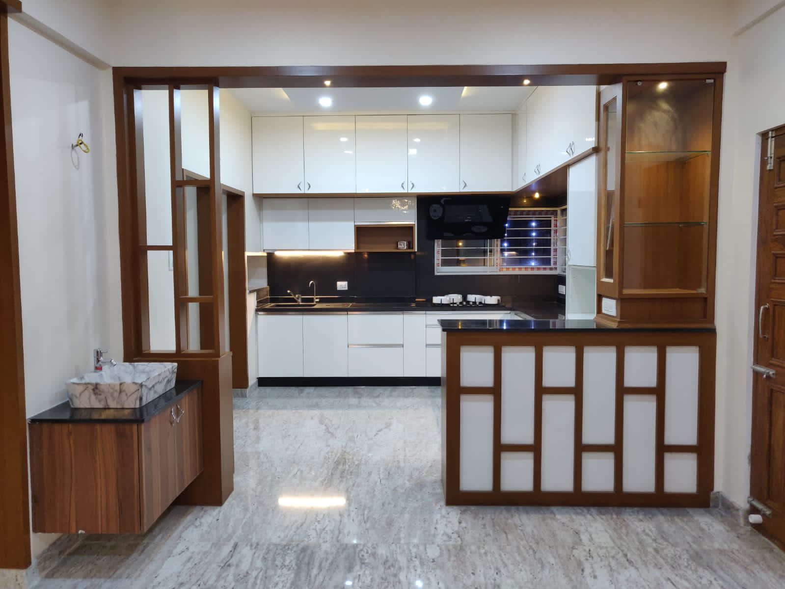 Modern kitchen interior design by Olangana in Bangalore, featuring sleek cream and taupe handleless cabinets, a light speckled countertop, and a black built-in hob with a slanted chimney.