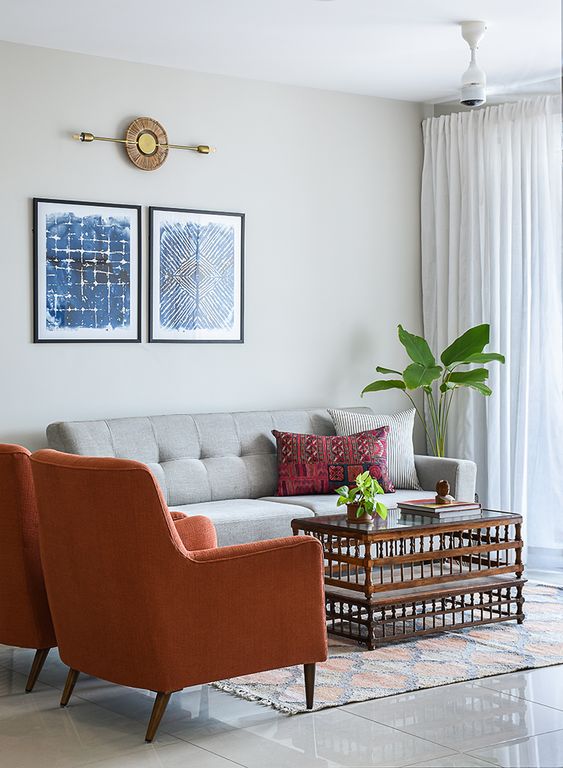 Modern living room interior design by Olangana in Bangalore, featuring a grey tufted sofa, burnt orange armchairs, blue abstract art, and a unique wooden coffee table.