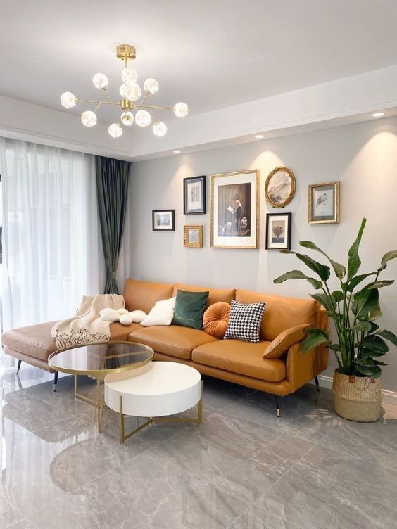 Modern living room interior design by Olangana in Bangalore, featuring a warm tan leather sectional sofa, stylish nesting coffee tables, a curated gallery wall, and a Sputnik chandelier.
