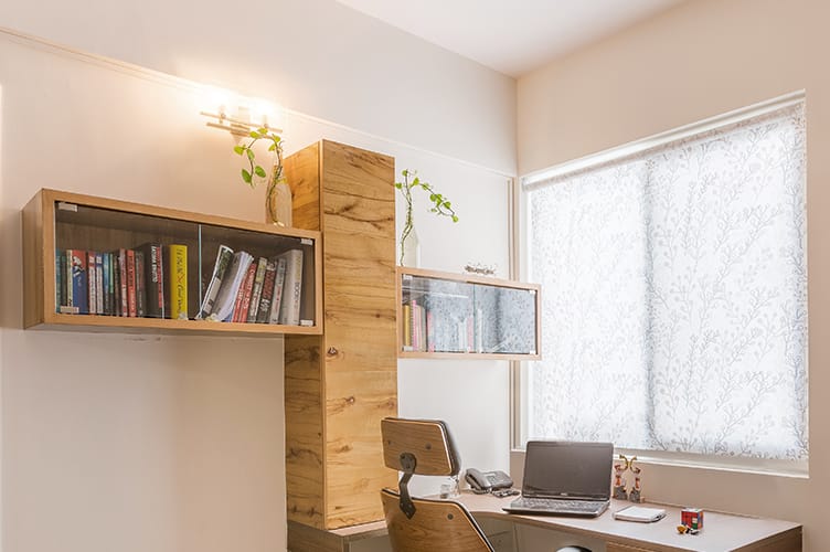 Elegant home office interior design by Olangana in Bangalore, featuring bespoke light wood shelves with glass fronts, a matching desk, ergonomic chair, and delicate patterned window blinds, illuminated by warm wall sconces.