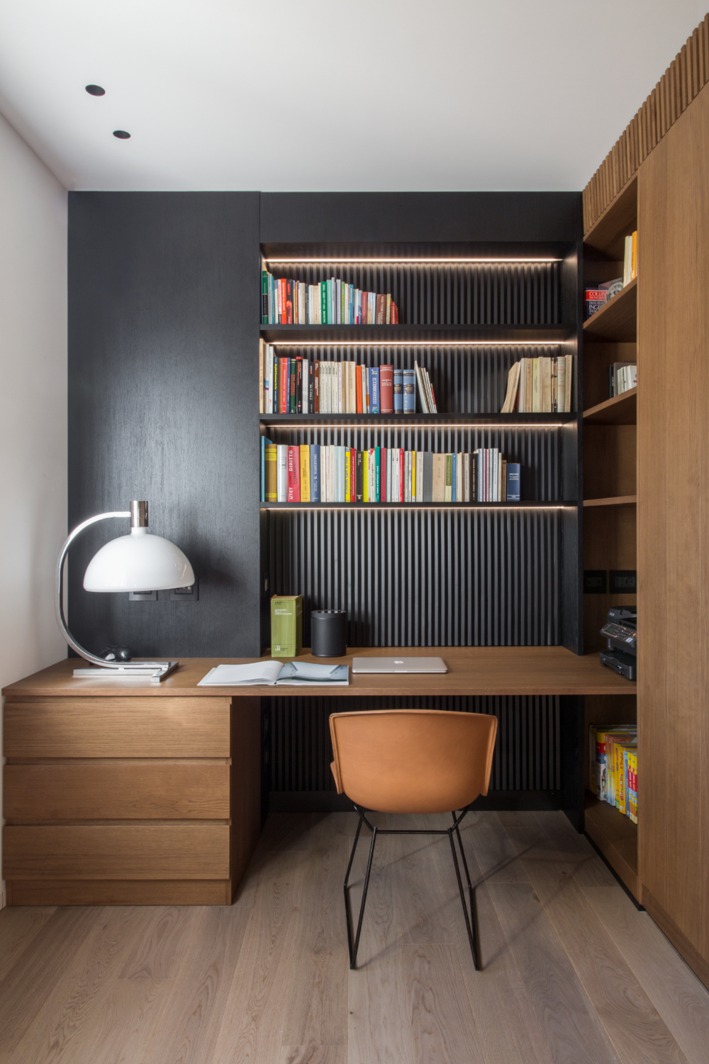 A sophisticated home office interior design by Olangana in Bangalore, featuring a custom light wood desk with drawers, integrated black ribbed bookshelves illuminated by warm LED strips, and a modern tan leather chair. This functional and elegant space showcases premium interior design.
