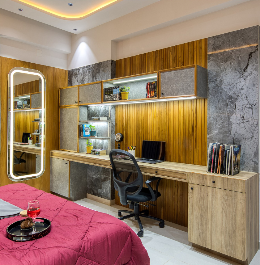 A contemporary bedroom interior design by Olangana in Bangalore, featuring a sleek integrated study area with warm wood slat paneling, sophisticated grey marble-effect walls, and illuminated shelving. The design includes a long wooden desk, an ergonomic chair, and a modern backlit mirror, showcasing premier Interior Design in Bangalore.