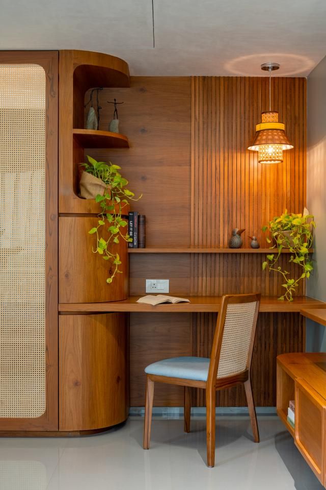 An elegant and functional home office or study area showcasing custom wooden built-ins, a rattan-backed chair with a light blue cushion, integrated shelving with lush green plants, and a stylish pendant lamp. This exquisite Interior Design by Olangana in Bangalore emphasizes warmth and productivity.