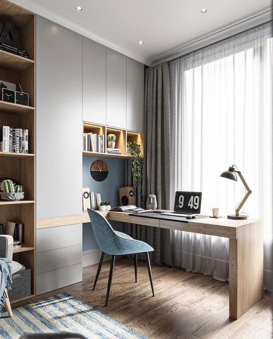 Elegant modern home office Interior Design by Olangana in Bangalore, featuring a light wood desk, a comfortable blue accent chair, sleek grey cabinetry, and integrated shelving with warm task lighting, bathed in natural light.
