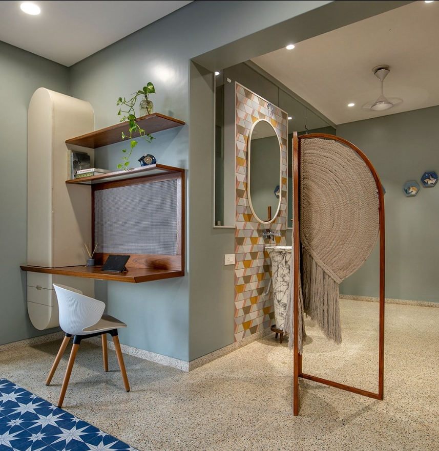 Contemporary Interior Design by Olangana in Bangalore, showcasing a stylish floating wooden desk with a modern white chair against sage green walls, complemented by a vanity area featuring patterned tiles, an oval mirror, and a unique marble pedestal sink, subtly divided by an elegant woven screen. This design emphasizes clever space utilization and sophisticated aesthetics.
