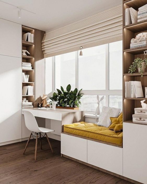 A modern and bright study nook designed by Olangana Interior Design, featuring a minimalist white desk, built-in wooden bookshelves with warm accent lighting, and a cozy yellow cushioned window seat, perfect for homes in Bangalore.