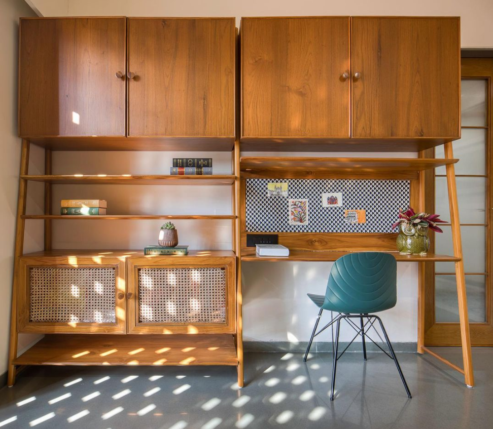 A sophisticated modular wooden shelving unit and integrated desk, designed by Olangana, featuring warm timber and elegant rattan accents with a modern teal chair. Exemplifying functional Interior Design in Bangalore, bathed in natural light.