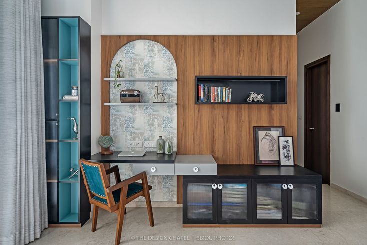 An elegant home office interior design by Olangana in Bangalore, featuring warm wooden wall paneling, a sleek black desk with light grey drawers, modern shelving with decorative items, and a striking teal upholstered chair. Adjacent is a tall black and blue bookshelf, showcasing Olangana's exquisite Interior Design.