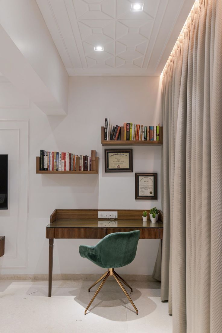 An elegant home office interior design by Olangana in Bangalore, showcasing a dark wood desk with a glass top, an emerald green velvet swivel chair, and floating wooden bookshelves. The space features modern recessed ceiling lights, intricate ceiling patterns, and soft neutral curtains, creating a sophisticated and functional workspace.