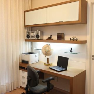 A modern and functional home office interior design by Olangana Design in Bangalore, featuring warm wooden built-in desk and floating shelves with bright under-shelf lighting, white frosted glass upper cabinets, an ergonomic office chair, and essential office equipment like a laptop and printer, set against soft white sheer curtains.