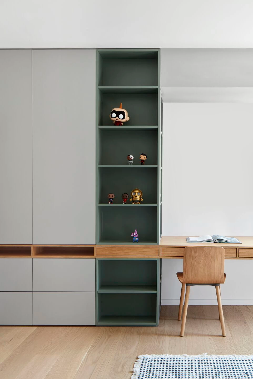 A modern and functional study area featuring bespoke light grey cabinets and integrated sage green open shelving displaying decorative figures. A natural wood desk and matching chair complement the light wooden flooring. This elegant Interior Design is a signature by Olangana in Bangalore.