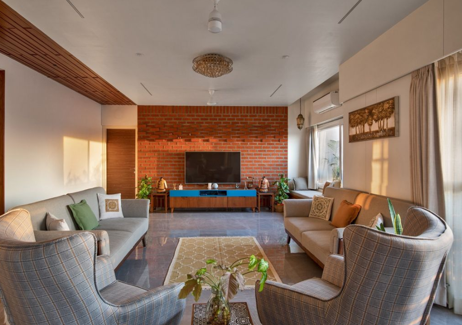 A warm and inviting living room interior design by Olangana in Bangalore, featuring a prominent exposed brick accent wall, comfortable grey and plaid sofas, a modern blue and wood media console, and a sophisticated blend of natural light and elegant fixtures. This Olangana Design embodies contemporary comfort.