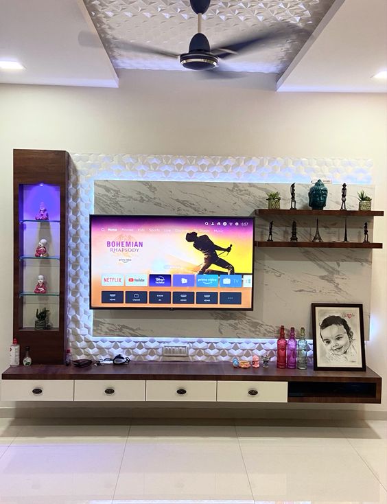 Modern living room interior design by Olangana in Bangalore featuring a sleek TV unit with white 3D textured paneling, integrated LED lighting, dark wood and white floating media console, and illuminated display shelves showcasing decorative elements.