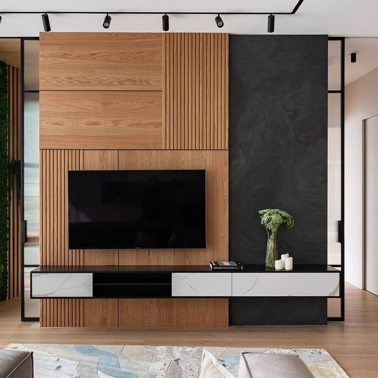 A sophisticated media wall by Olangana Interior Design in Bangalore, showcasing natural wood paneling, a dark textured accent wall, and a sleek floating TV unit with white marble-effect drawers, perfectly illuminated by modern track lighting.
