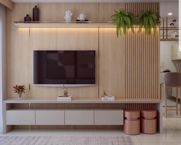A modern living room Interior Design by Olangana in Bangalore, featuring a TV wall with light wood paneling, a floating shelf with integrated lighting, decorative plants, and a sleek grey media console with accent stools.