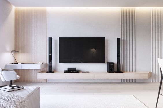 A modern minimalist living room by Olangana Design in Bangalore, featuring a wall-mounted TV, floating light wood media console, and sleek black speakers. The elegant Interior Design incorporates subtle cove lighting, textured wall panels, and a functional floating desk area, epitomizing sophisticated living spaces in Bangalore.