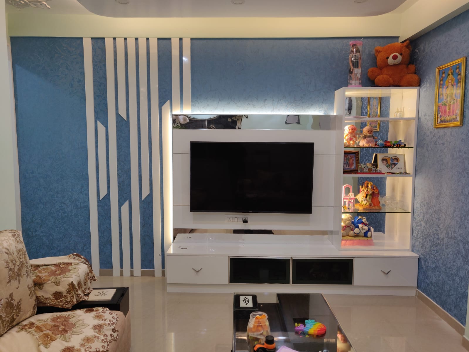 Modern living room Interior Design by Olangana in Bangalore, featuring a stylish white TV unit with integrated shelving against a blue textured wall with elegant white geometric paneling and soft accent lighting. This Olangana Design showcases sophisticated residential interior design.