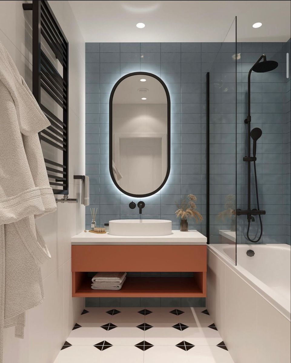 A modern bathroom interior design by Olangana in Bangalore, featuring dusty blue subway tiles, an oval backlit mirror, a vibrant orange floating vanity with a white vessel sink, and sleek black fixtures. The geometric black and white floor tiles add a distinctive touch to this elegant space, showcasing Olangana Design expertise.
