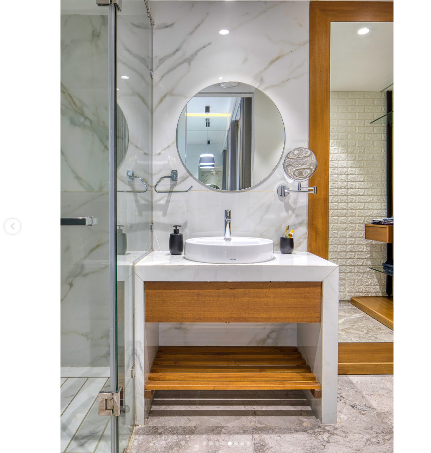 An elegant bathroom interior design by Olangana in Bangalore, showcasing a modern vanity with white marble countertop and backsplash, a round illuminated mirror, a chic vessel sink, and warm wooden cabinetry with an open slatted shelf. A glass shower enclosure is visible on the left.