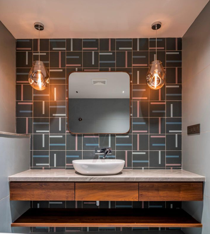 Elegant bathroom interior design by Olangana in Bangalore, showcasing a floating wooden vanity with a marble countertop, a pristine white vessel sink, and a modern rounded mirror. The accent wall features stylish dark grey geometric tiles with pink, blue, and white patterns, illuminated by two warm, teardrop-shaped pendant lights.