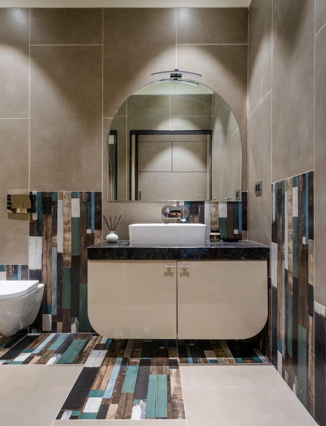 Elegant powder room Interior Design by Olangana in Bangalore, featuring a floating cream vanity, dark countertop, white basin, and an arched mirror with accent lighting. The space is enhanced by unique patterned wall and floor tiles resembling distressed wood planks in a vibrant mix of blues, greens, and earthy tones, offering a sophisticated and modern aesthetic crafted by Olangana Design.