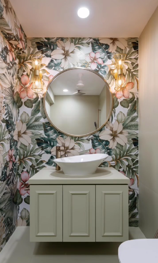 Exquisite powder room Interior Design by Olangana in Bangalore, featuring vibrant tropical floral wallpaper, a chic light green floating vanity, a round gold-framed mirror, and elegant gold sconces that create a luxurious and inviting ambiance.