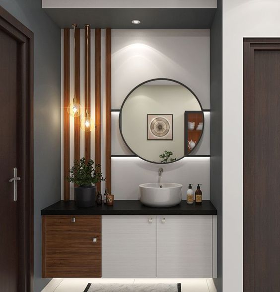 An elegant modern powder room interior design by Olangana in Bangalore, featuring a round black-framed mirror, a crisp white vessel sink, a sleek dark countertop, and a floating vanity with a mix of white and warm wooden finishes, accented by vertical wooden slat paneling and stylish pendant lights, all reflecting premium Interior Design.